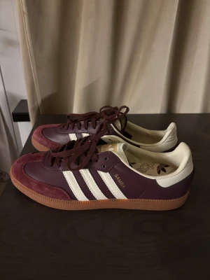 Adidas Samba vinröda sneakers - Så himla CHIC Adidas Samba sneakers i vinrött och vitt med klassiska tre ränder på sidan. Ovandelen är i skinn och mocka, med brun gummisula och snörning. Insidan är vit och har Adidas-logga på innersulan. Perfekta för dig som vill ha en retro och stilren look. Helt nya och oanvända, har också vita skosnören som följde med, och följer med vid köp.  