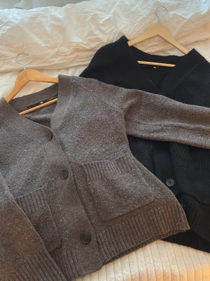 Grå och svart kofta från 157 - Två mysiga koftor från 157 i storlek S, en i grått och en i svart. Båda har ribbade detaljer vid fickor och nederkant, samt stora knappar framtill. Koftorna är stickade och har en loose passform med långa ärmar och djup v-ringning.
