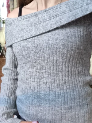 Grå ribbad offshoulder stickad tröja - Säljer en snygg grå stickad tröja med ribbad struktur och bred offshoulder-krage. Tröjan har lång ärm och är perfekt för dig som gillar en chill och trendig look. Passar dig som vill ha något mjukt och bekvämt med lite extra stil.