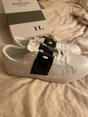 Valentino Rockstud Untitled sneakers - Säljer ett par vita Valentino Rockstud Untitled sneakers med svart läderpanel och ikoniska pyramidnitar. Skorna har rund tå, platt sula och snörning. Märket syns på plösen och innersulan. Riktigt snygga och sticker ut med sina svarta detaljer och rockiga vibe. De var min brors men säljer då de tyvärr var för små. De är i storlek 43. Det finns tyvärr inget kvitto på dom då jag köpte dom här på plock för 2500 💗