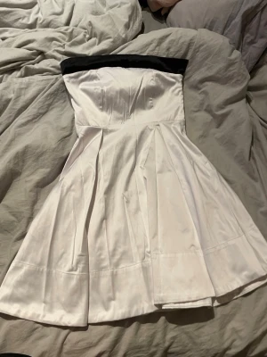 Vit axelbandslös klänning med rosett - Supersöt axelbandslös vit klänning med svart kant upptill och en stor svart rosett framtill. Klänningen har en utsvängd kjol och är gjord i ett glansigt material som känns lyxigt. Perfekt för dig som vill sticka ut med en klassisk men ändå lekfull look.