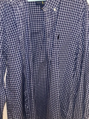 Blårutig skjorta från Ralph Lauren - Klassisk blårutig skjorta från Ralph Lauren med vit rutmönster, knappkrage och bröstficka. Skjortan har långa ärmar och knappar framtill. Perfekt för dig som gillar tidlös stil och vill ha något som funkar till det mesta.