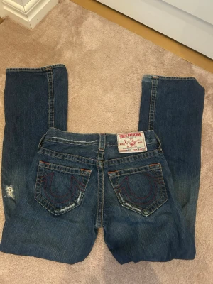True Religion Jeans bootcut  - Säljer ett par klassiska blå jeans från True Religion med tydliga kontrastsömmar i orange och rött. Jeansen har utsvängda ben, ikoniska bakfickor med broderad logga och slitna detaljer på benen. Tillverkade i robust denim med cool vintagekänsla.