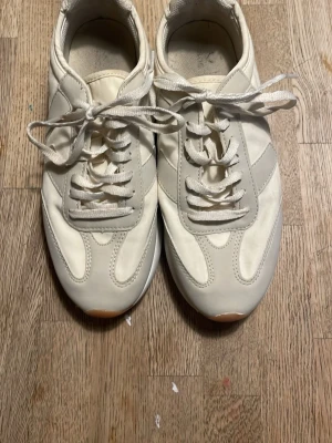 Vita sneakers från Stadium strl 36 - Säljer ett par vita sneakers från Stadium i storlek 36. Skorna har snörning, rund tå och en platt sula med orange mönstrad undersida. Ovandelen är i syntetmaterial och textil, med stilrena paneler och klassisk sneaker-look.