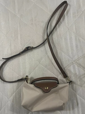 Beige axelväska med bruna detaljer - Snygg beige axelväska med justerbart brunt axelband och lock i brunt skinn med guldfärgad knapp. Perfekt storlek för det viktigaste och stilren design som passar till många outfits.