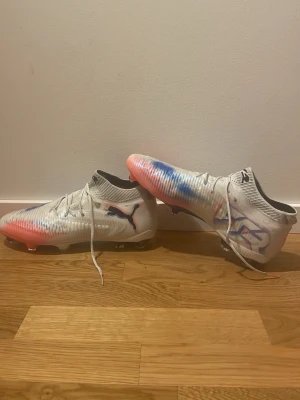 Puma future - Säljer ett par grymma vita Puma fotbollsskor med coola blå och röda färgstänk. Skorna har snörning, låg profil och stickad strumpliknande öppning för extra komfort. Samt de är aldrig använda!  Sulan är designad för bra grepp på planen och har svarta och orange detaljer. Perfekta för dig som vill sticka ut på matchen.