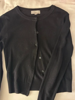 Svart kofta från H&M - En klassisk svart kofta från H&M med rund halsringning och knappar framtill. Koftan har långa ärmar och ribbade muddar vid ärmslut och nederkant.