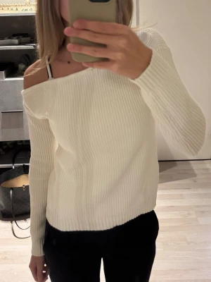 Vit ribbad offshoulder stickad tröja - Säljer en vit stickad tröja med ribbad struktur och offshoulder-design. Tröjan har lång ärm och en snygg, lite loose passform som ger en avslappnad vibe. Användes 1 gång.