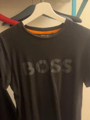 Svart BOSS t-shirt med tryck - Snygg svart t-shirt från BOSS i storlek M. T-shirten har ett stort BOSS-tryck på bröstet och klassisk rund hals. Passformen är normal och materialet känns mjukt och bekvämt, perfekt för en chill och stilren look.