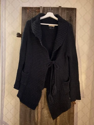 Svart stickad kofta Odd Molly - Mysig svart stickad kofta från Odd Molly med grov ribbad krage, knytband i midjan och två fickor fram. Koftan har lång ärm och är tillverkad i en mjuk mix av akryl, alpacka och ull. Perfekt för lager på lager under kyliga dagar eller svala sommarkvällar.