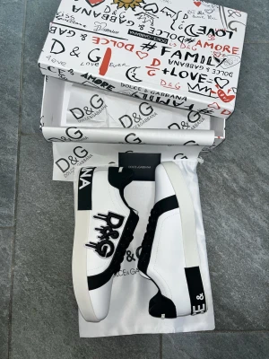 Dolce & Gabbana vita och svarta sneakers - Snygga vita och svarta sneakers från Dolce & Gabbana med stora D&G-loggor på sidan och hälen. Skorna har svarta snören, rund tå och detaljer i svart skinn. Tjock vit sula och cool kontrast mellan färgerna. Perfekta för dig som vill sticka ut med en exklusiv look.