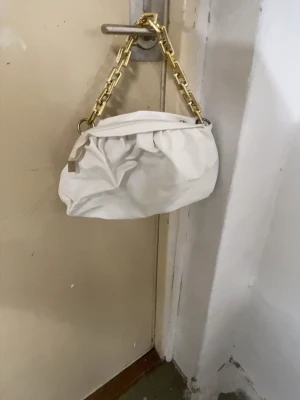 Vit axelväska med guldkedja - Säljer en snygg vit axelväska med en chunky guldfärgad kedja som axelrem. Väskan har en mjuk, puffig form och stängs med dragkedja. Perfekt för dig som vill ha en trendig accessoar som sticker ut.