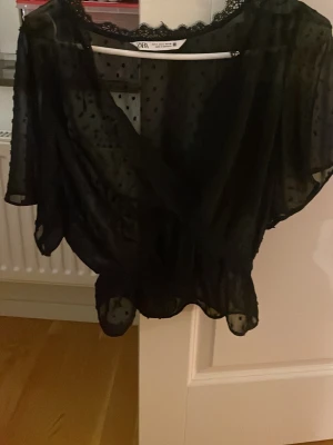Svart prickig omlottblus från Zara - Säljer en svart omlottblus från Zara med transparent tyg och små prickar. Blusen har spetsdetaljer vid halsringningen och korta, lösa ärmar. Den är i storlek M!