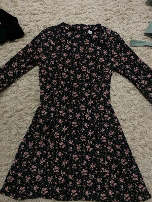 Blommig svart klänning från H&M Divided - Supersöt svart kortklänning från H&M Divided med små rosa och blå blommor. Klänningen har långärmade ärmar och en lätt utställd kjol. Perfekt för dig som gillar romantiska och feminina plagg. Går också jätte bra på Barn 