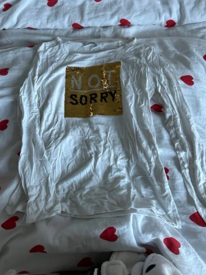 Vit långärmad t-shirt med paljetter - Vit långärmad t-shirt från C i storlek 134/140. Framsidan har en stor fyrkant med vändbara paljetter som skiftar mellan guld med texten 'NOT SORRY' och färgglada paljetter med texten 'SORRY'. Mjuk och skön bomullskänsla, perfekt för dig som gillar statement-plagg.