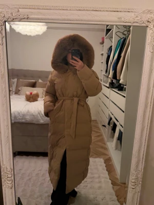 Lång beige dunjacka med pälshuva - Säljer en lång beige dunjacka med stor huva kantad av päls. Jackan har bälte i midjan för snygg passform och är perfekt för kalla dagar. Den är quiltad och har långa ärmar samt en slits nertill för extra rörelsefrihet.