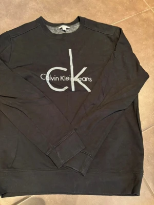 Svart sweatshirt från Calvin Klein Jeans - Svart sweatshirt från Calvin Klein Jeans med stor CK-logga framtill. Tröjan har rund halsringning, långa ärmar och ribbade muddar vid ärmslut och nederkant. Tillverkad i 100% bomull för en skön och avslappnad känsla.