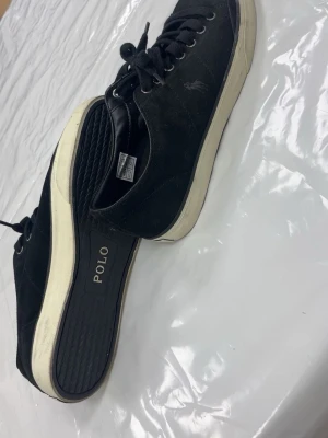 Svarta sneakers från Polo Ralph Lauren - Snygga svarta sneakers från Polo Ralph Lauren med vit sula och klassisk POLO-logga på hälen. Skorna har snörning och är gjorda i mocka för en stilren look. Perfekta till jeans eller shorts för en clean och enkel stil. Kan få tvättade!