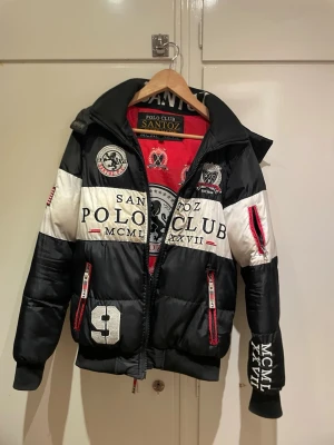 Svart och vit pufferjacka SANTOZ Polo Club - Säljer en fet pufferjacka från SANTOZ Polo Club i svart och vitt med stora broderade loggor och patchar. Jackan har röd insida, dragkedja framtill, flera fickor med röda detaljer och coola tryck på både fram- och baksida. Perfekt för dig som vill sticka ut i vinter! Hål i höger ficka och litet hål över vänster ficka men båda fickorna går att använda utmärkt