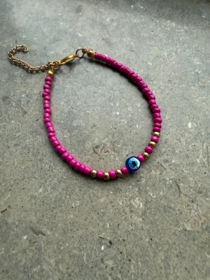 Rosa pärlarmband  - Säljer ett handgjort armband med små rosa pärlor, guldfärgade detaljer och en blå evil eye-pärla. Armbandet har justerbar kedja i guldton och passar perfekt för att ge din outfit lite extra färg och personlighet.
