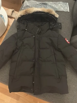Svart dunjacka från Canada Goose - Svart dunjacka från Canada Goose med pälskantad huva och klassisk logga på ärmen. Jackan har dragkedja och knappar framtill, samt flera praktiska fickor. Perfekt för kalla vinterdagar och har en riktigt schysst oversized look.