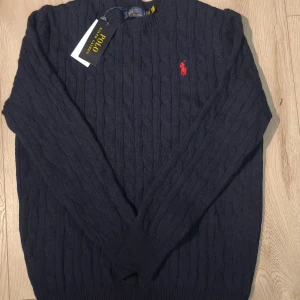 Mörkblå Ralph Lauren Knit M - Stickad Ralph Lauren-tröja i mörkblått med den klassiska Polo-loggan på bröstet. Tillverkad i 100 % mjuk bomull som sitter bekvämt och håller formen fint. Oanvänd och i mycket fint skick. Storlek M med längd 65 cm och bredd 48,5 cm. Hör gärna av dig vid frågor.