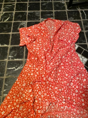 Röd blommig omlottklänning - Superfin röd omlottklänning med vita blommor och korta ärmar. Klänningen har knytband i midjan och volangdetaljer nertill. Perfekt för varma dagar och ger en riktigt somrig vibe. Tunn och luftig, troligtvis i polyester eller bomull.