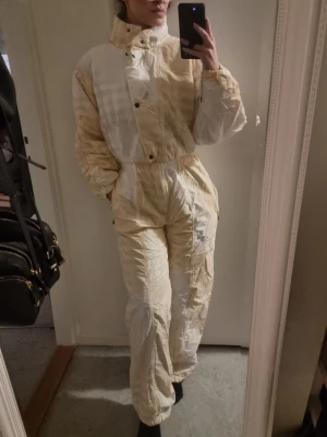 Vintage täckbyxor från Lutha - Retro täckbyxor från Lutha i ljusbeige och vitt med diskret rutigt mönster. Byxorna har hög midja, resår i midjan och benslut samt dragkedja och knappar framtill. Perfekta för vintern och riktigt sköna att ha på sig.