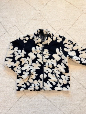 Svart och vit teddyjacka från HUF - Säljer en fet teddyjacka från HUF med svart botten och vita, abstrakta mönster. Jackan har dragkedja framtill, två fickor med dragkedja och ett camo-mönstrat innerfoder. Perfekt för dig som vill sticka ut med en unik look.