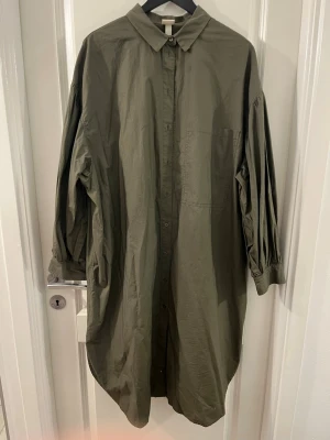 Olivgrön skjorta från H&M, storlek M - Säljer en olivgrön skjorta från H&M i storlek M. Skjortan har en loose passform, klassisk krage, långa ärmar med knapp vid manschetten och en bröstficka. Materialet är lätt bomull och skjortan har knappar hela vägen framtill.