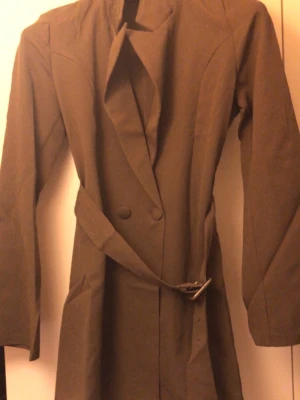 Brun trenchcoat med bälte - Snygg brun trenchcoat med klassisk krage och bälte i midjan. Jackan har dubbelknäppning framtill och långa ärmar. Perfekt för dig som vill ha en stilren och tidlös look. Materialet känns som bomull eller polyester.