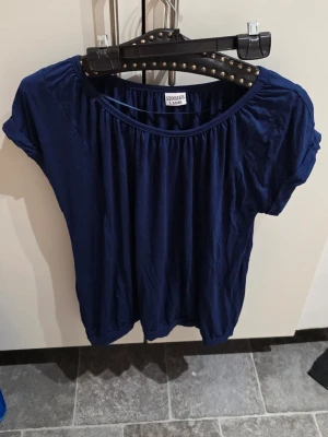 Mörkblå topp från Stories, storlek L - Mjuk och flowy mörkblå topp från Stories i storlek L. Tillverkad i lyocell och elastan för en skön stretchig känsla. Toppen har rund halsringning, korta ärmar och lätt puffig passform med rynkade detaljer vid halsen och nederkanten.