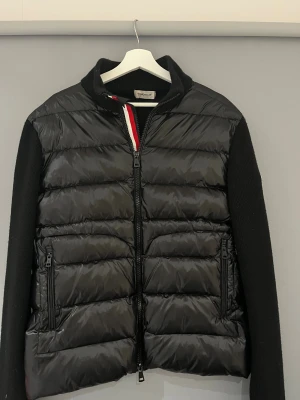 Moncler cardigan - Säljer en svart Cardigan från Moncler med stickade ärmar och quiltad front. Jackan har dragkedja framtill, två fickor med dragkedja och en liten Moncler-logga på ärmen. Jackan är storlek XXL men sitter som L/XL. Jackan är även i väldigt bra skick, det ända är att moncler lappen i nacken släppt lite (se sista bild) men förövrigt en riktig go jacka. Hör av er vid minsta funderingar! (Osäker om jackan är 0rginal)