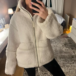 Teddyjacka vit med dragkedja - Mysig och fluffig vit teddyjacka med hög krage och stora fickor framtill. Jackan har en rejäl dragkedja och är perfekt för kyliga dagar. Modellen är loose fit, går över rumpan och har långa ärmar för extra komfort. 