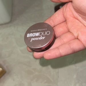 Makeup Mekka Brow Duo Powder - Säljer en Brow Duo Powder från Makeup Mekka i en rund, rosa och svart dosa. Innehåller två matta nyanser i ljusbrun och mellanbrun för att fylla och forma brynen. Perfekt för att skapa naturliga eller markerade bryn. Smidig att ta med i väskan.