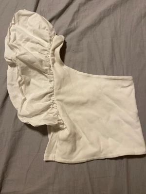 Vit oneshoulder topp med volangärm - Säljer en vit oneshoulder topp med en stor, rynkad volangärm på ena sidan. Toppen är croppad och är i storlek S