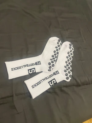 FS FootballSocks vita träningsstrumpor - Säljer ett par vita FS FootballSocks med svarta detaljer och grepp på undersidan. Strumporna har ribbad design och tryckt logga längs skaftet. Perfekta för fotboll eller annan träning där du vill ha extra grepp i skorna.