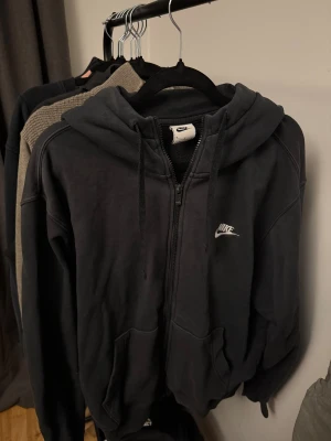 Svart zip-hoodie från Nike - Svart hoodie från Nike med dragkedja och broderad vit logga på bröstet. Klassisk modell med huva och dragsnören samt två stora fickor framtill. Mjuk bomullsmix som är skön att bära. Perfekt för en chill och sportig stil.