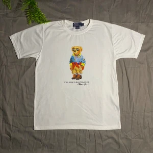 🐻Vit Polo Bear t-shirt från Ralph Lauren🐻 - Vit t-shirt från Polo Ralph Lauren med ikoniskt Polo Bear-tryck på bröstet. T-shirten har rund hals, korta ärmar och är gjord i mjuk bomull. Trycket visar en cool björn med solglasögon och streetwear-outfit. Perfekt för dig som gillar klassisk stil med en twist.