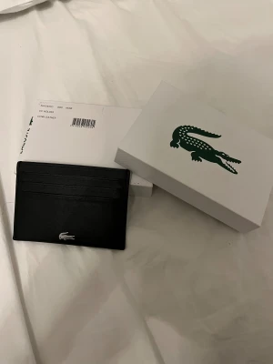 Lacoste plånbok - Stilren svart korthållare i äkta läder från Lacoste. Platt och smidig design med tre kortfack på varje sida och en silverfärgad Lacoste-logga framtill. Perfekt för dig som vill ha ett minimalistiskt och snyggt sätt att bära dina kort.