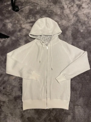 Vit hoodie med dragkedja - Snygg vit hoodie med broderad logga på fickan och mönstrat foder i huvan. Tröjan har hel dragkedja, snörning i huvan och ribbade muddar. Materialet är stickad bomull som känns mjuk och lyxig. Perfekt för dig som gillar clean och exklusiv streetwear.