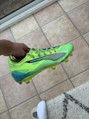 Puma limegröna fotbollsskor - Säljer ett par snabba Puma fotbollsskor i limegrönt med blå detaljer och svart logga på sidan. Skorna har snörning, platt sula med fasta dobbar och är tillverkade i syntetmaterial för lätt känsla på planen. Perfekta för dig som vill sticka ut på matchen!
