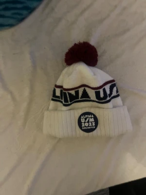 USM Alpina 2025 – Åre/Duved mössa - Säljer en vit vintermössa från USM Alpina 2025 Åre/Duved. Mössan har en fin röd tofs på toppen och en stilren design med blå och röda detaljer runtom. Perfekt minnespryl för dig som gillar alpint eller vill hålla dig varm i skidbacken. Mjukt och skönt material som sitter bra i vinterkylan.