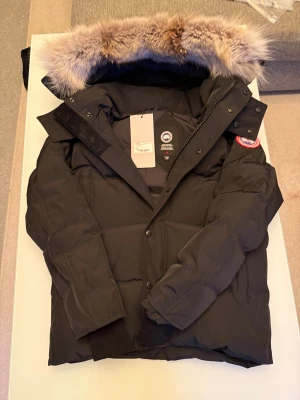 Svart Wyndham Parka från Canada Goose - Svart Wyndham Parka från Canada Goose med ikonisk patch på ärmen och fluffig päls på huvan. Jackan är dunfodrad, har dragkedja och knappar framtill samt ribbade muddar. Perfekt för kalla vinterdagar och har en clean, klassisk look.