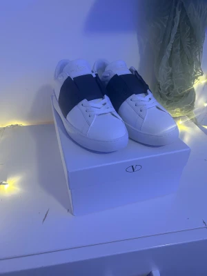 Valentino Garavani Open sneakers vit/blå - Snygga Valentino Garavani Open sneakers i vitt skinn med bred blå läderdetalj över snörningen. Klassisk rund tå, platt sula och vita snören. Diskret logga på plösen och coola nitar baktill på hälen. Perfekt för dig som gillar lyxiga och stilrena sneakers.