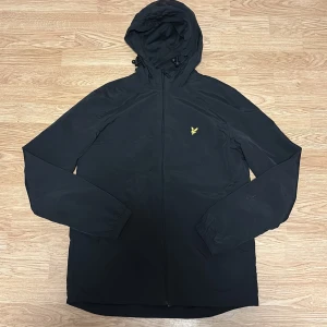 Svart vindjacka från Lyle & Scott - Svart vindjacka från Lyle & Scott helt ny med huva och gul logotyp på bröstet. Jackan har dragkedja framtill, två sidofickor med knappstängning och justerbar huva med snörning. Perfekt för blåsiga dagar och regniga dagar. Hör av er vid intresse 