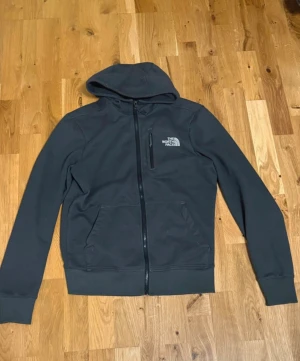 Grå/svart fleecejacka från The North Face - Svart fleecejacka med huva från The North Face. Jackan har hel dragkedja framtill, två sidofickor och en bröstficka med dragkedja. Mjuk insida i fleece och klassisk logga på bröstet. 