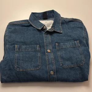 Overshirt i denim från H&M - Snygg blå denim overshirt från H&M med klassisk krage, två stora bröstfickor och knappar framtill. Tillverkad av ekologisk och återvunnen bomull. Perfekt lager-på-lager-plagg med lång ärm och avslappnad passform.