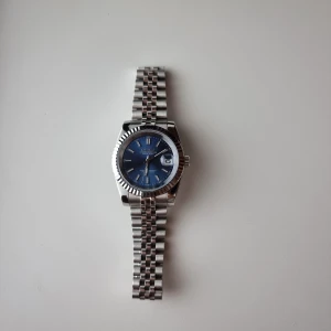 Seiko herrklocka med blå urtavla - Säljer min Saiko mod-klocka som är byggd i stil med en Datejust. Den har en elegant mörkblå/marinblå urtavla och ett klassiskt Jubilee-armband som ger en riktigt tidlös känsla. Klockan är 39 mm i diameter och har självlysande visare samt tim- och minutmarkeringar, vilket gör den lätt att läsa även i mörker. Den har burits i cirka två veckor och är därför i princip som ny, med endast något enstaka litet spår av användning. Seiko NH35A urverk. Klockan levereras med en box och fungerar felfritt.