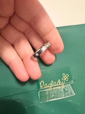 Snygg silverring med stenar Raglady - Stilren och cool silverfärgad ring från Raglady by Tara. Ringen är rund och har en platt yta med tre infattade klara stenar som ger extra bling. Perfekt för dig som gillar minimalistisk design med en touch av lyx. Kommer i original ask.